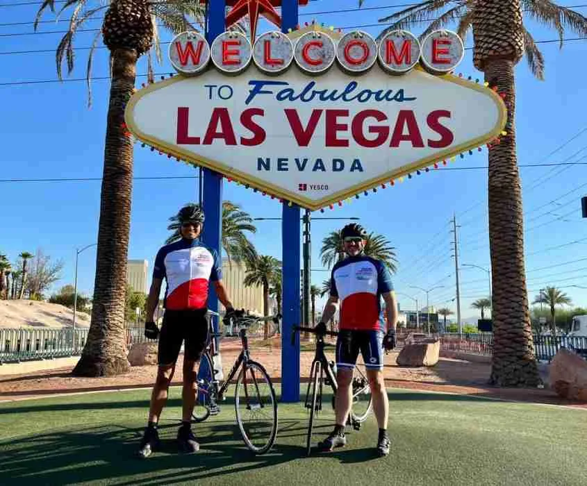 Two RAFA Rides cyclists standing with their bikes in front of a big sign saying "Welcome to fabulous Las Vegas" with palm trees either side.
